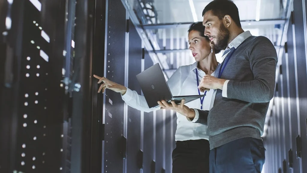 A man and a woman discussing something at a laptop next to the control panel